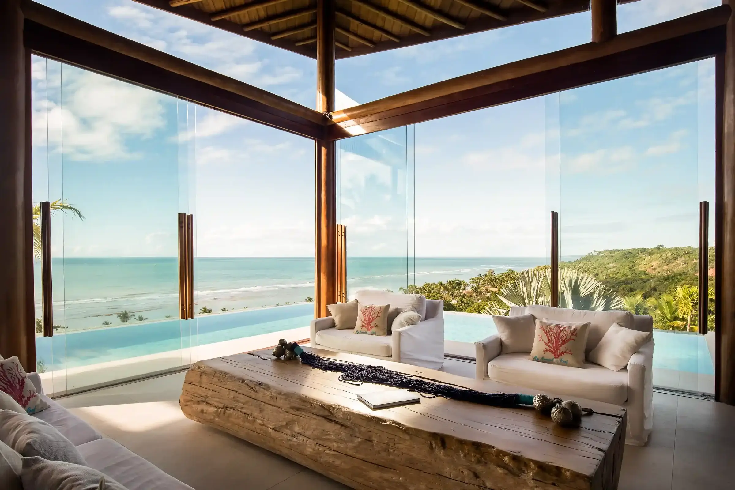 Glass-walled living room with a log table and light sofa, sea and pool behind