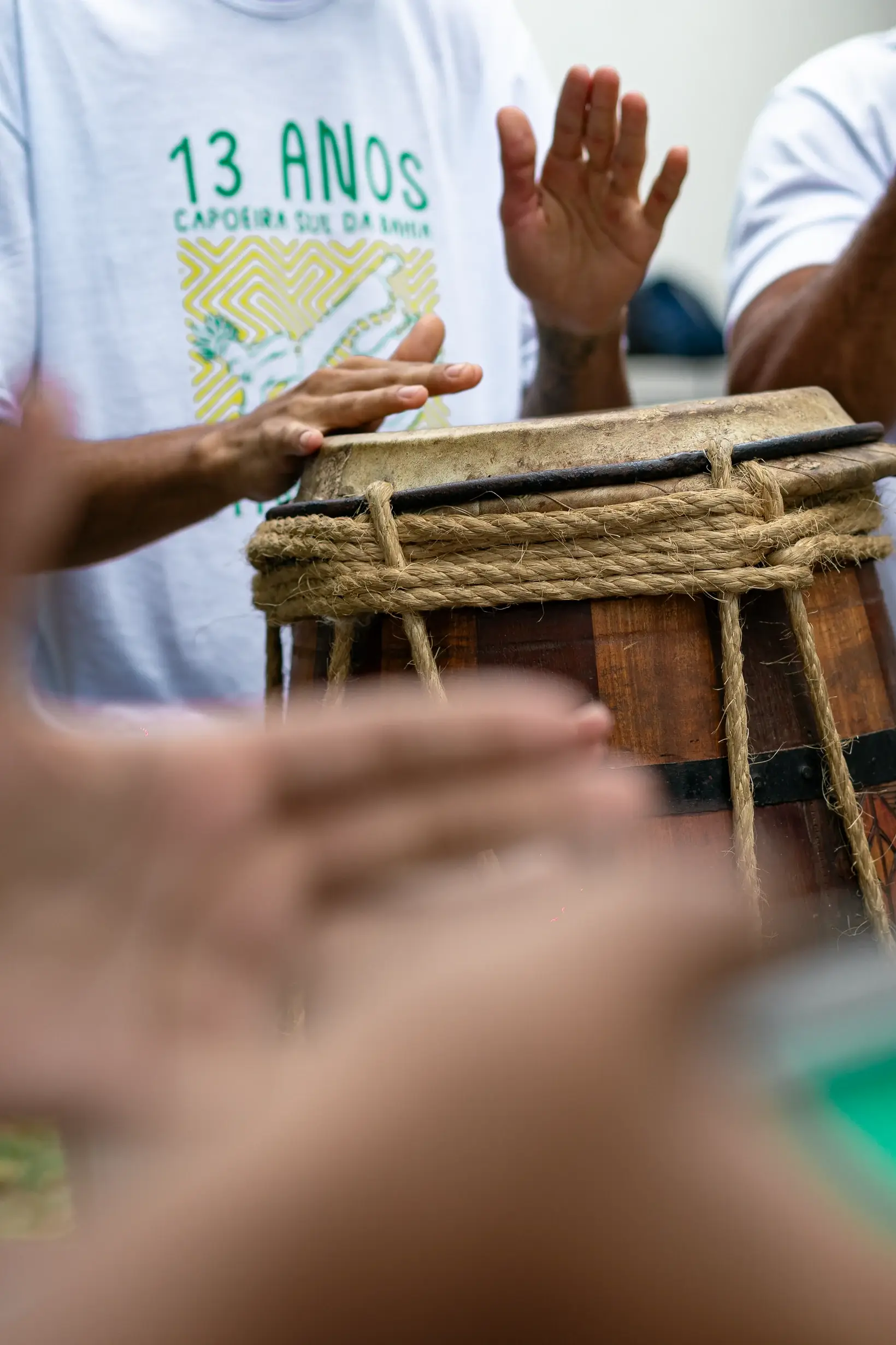 Mãos tocando atabaque com corda trançada, camiseta dos 13 anos do grupo ao fundo