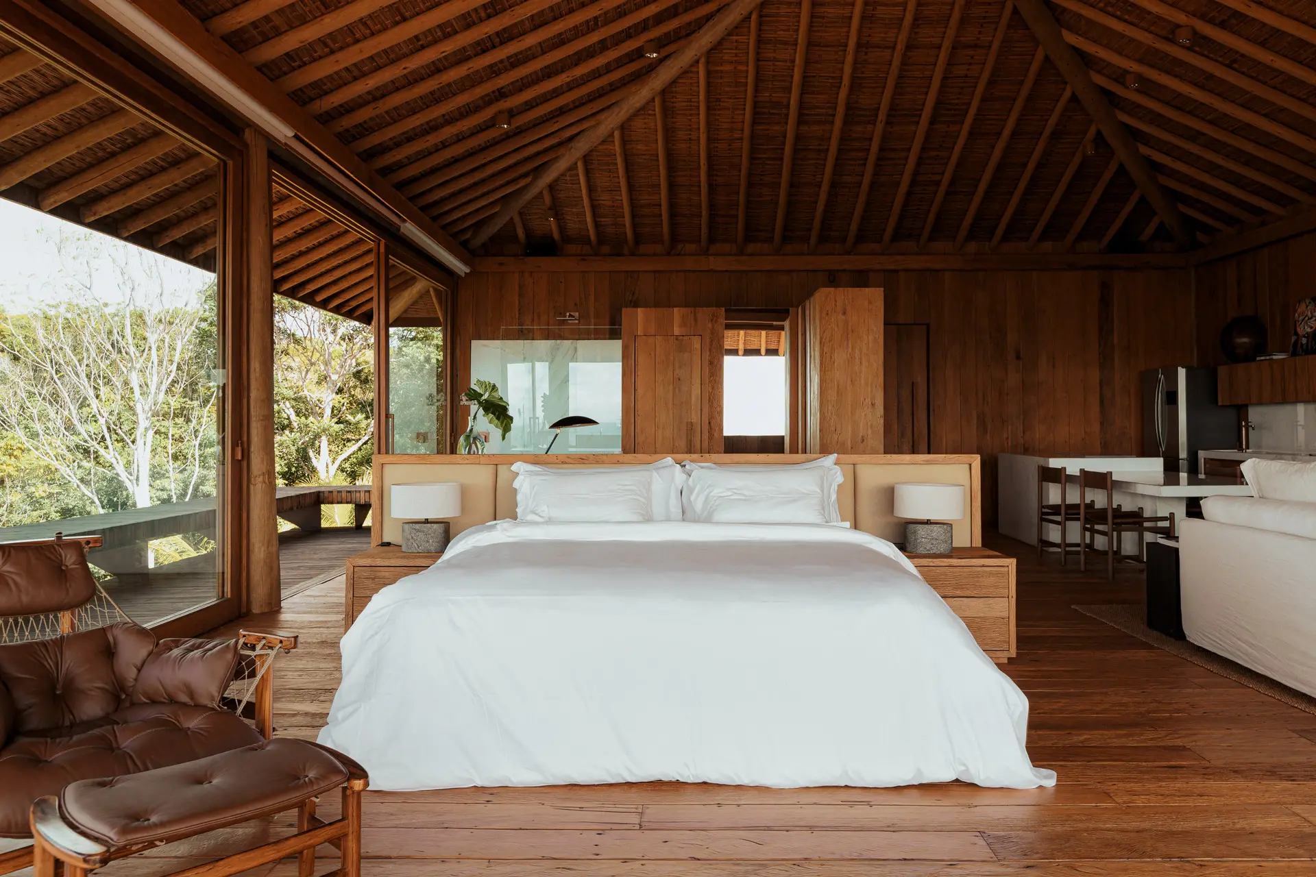 Quarto com cama de casal, forro de madeira em duas águas e poltrona de couro, aberto para a mata