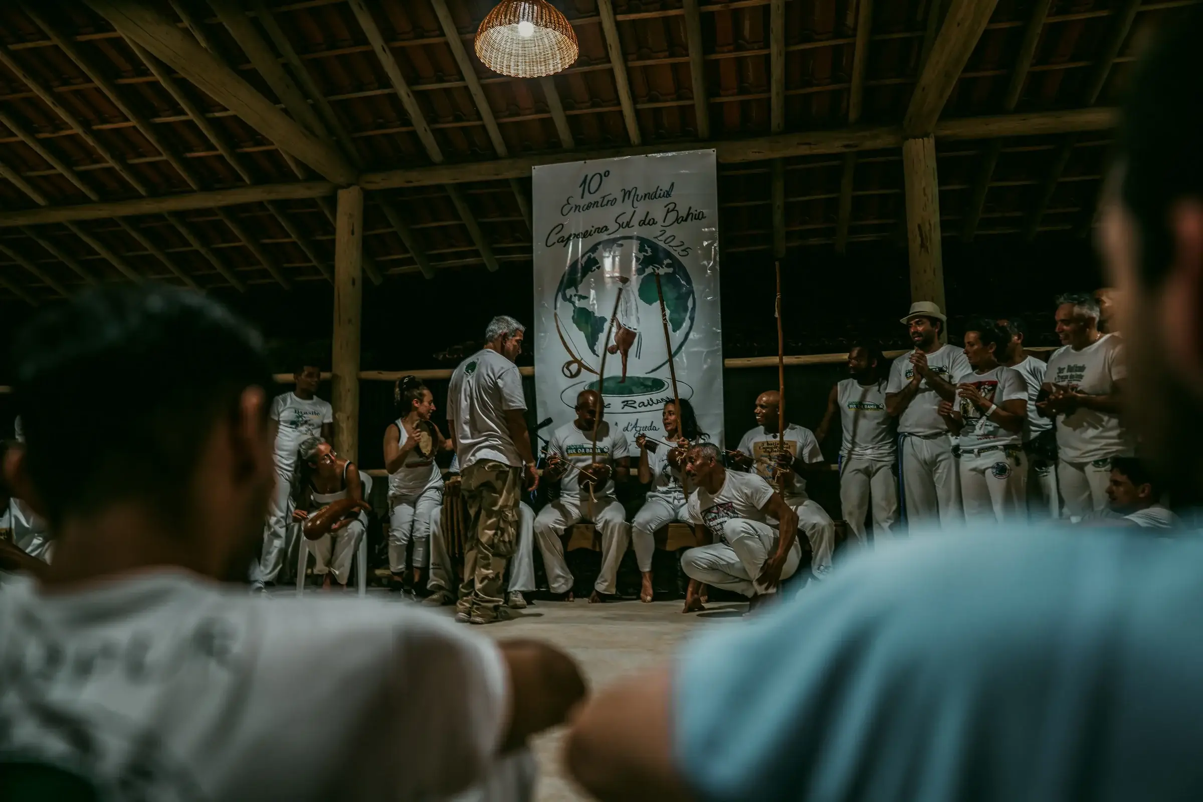 Roda de capoeira em salão com faixa do grupo ao fundo, capoeiristas de branco