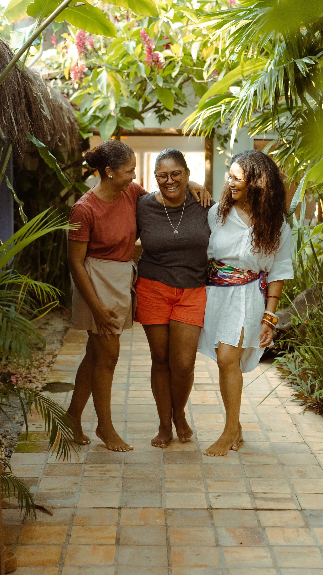 Três mulheres abraçadas sorrindo num caminho de pedra entre plantas