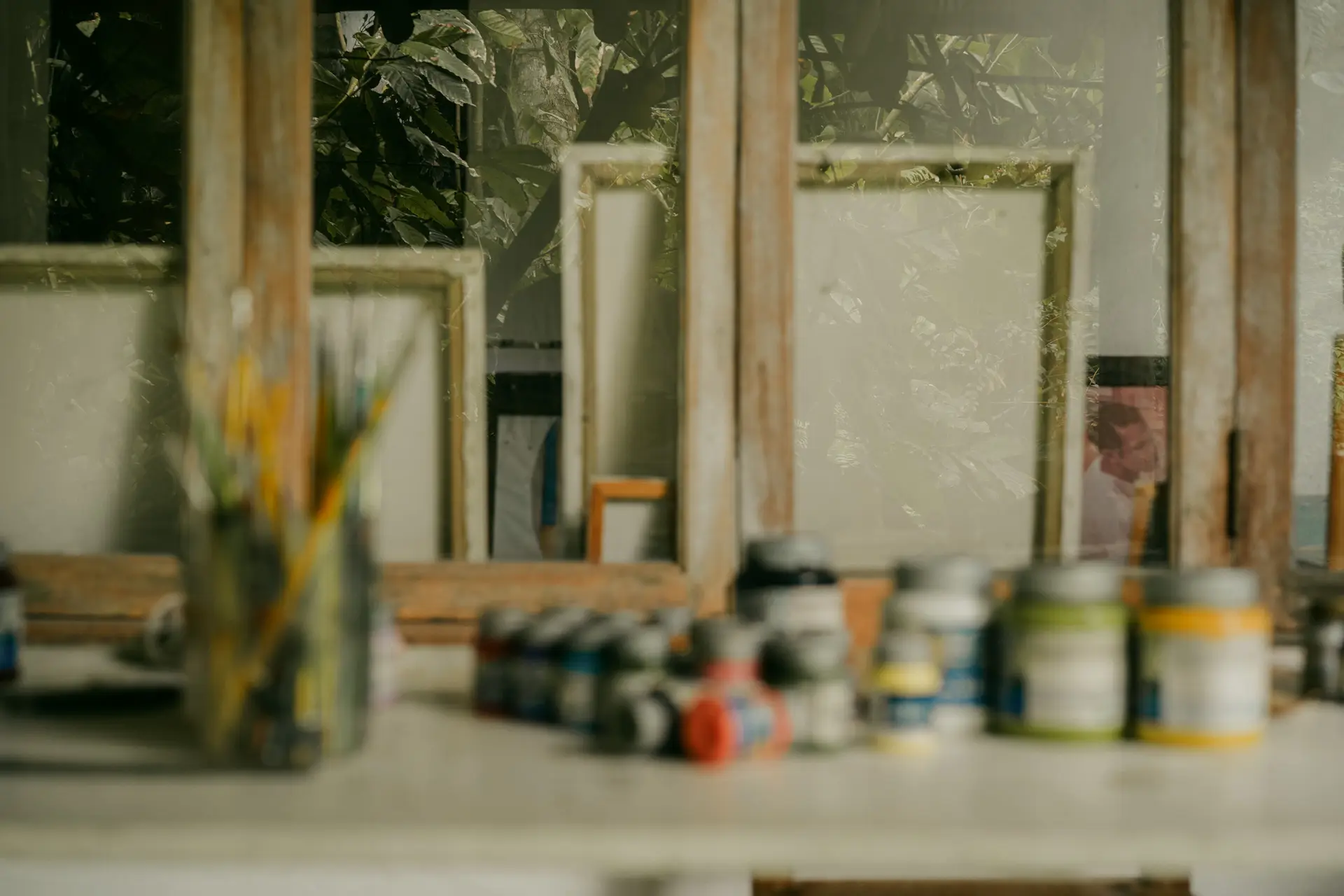 Potes de tinta e pincéis sobre a bancada do ateliê, telas ao fundo