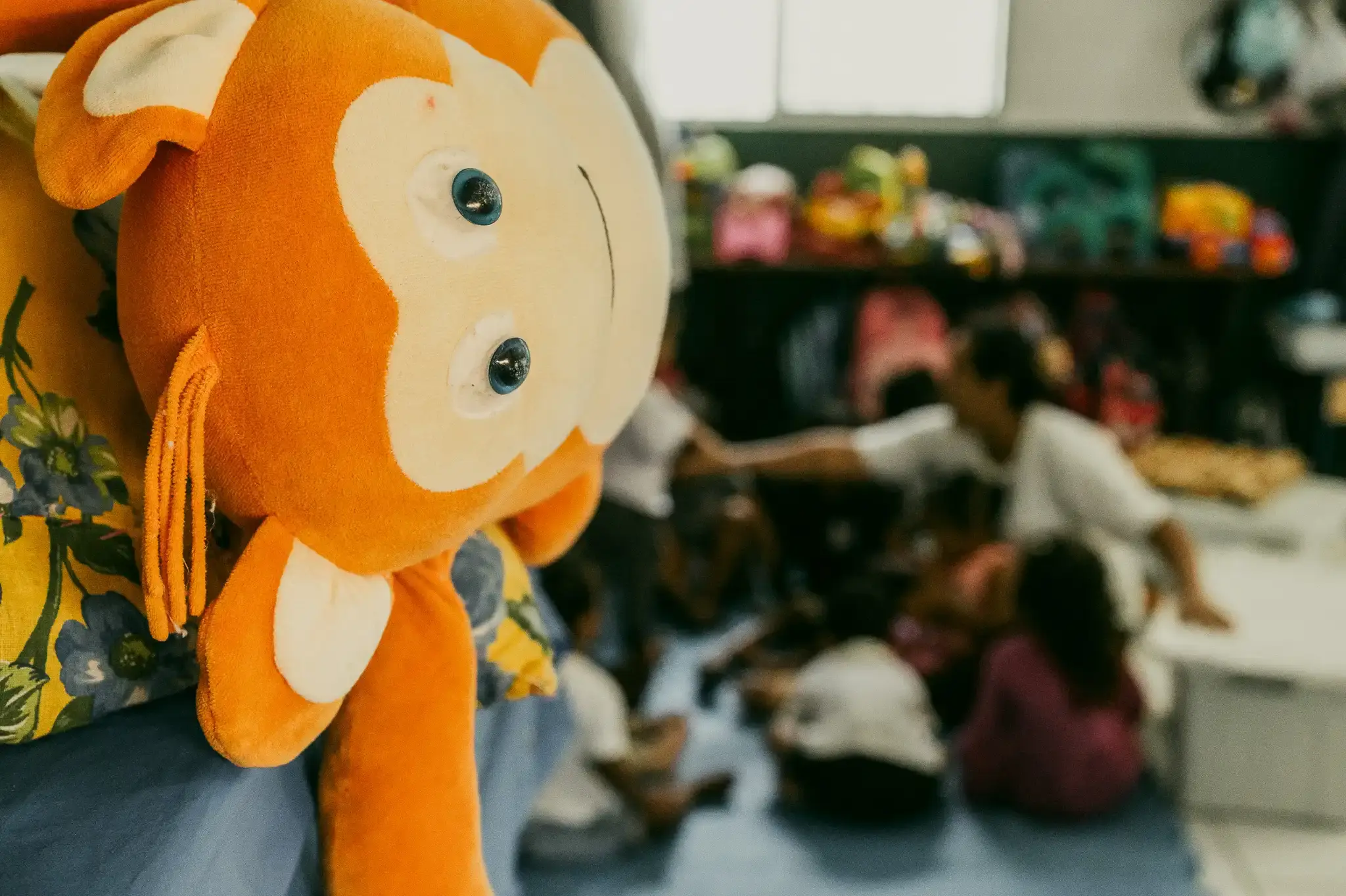 Boneco de pelúcia laranja em primeiro plano e crianças brincando na sala ao fundo