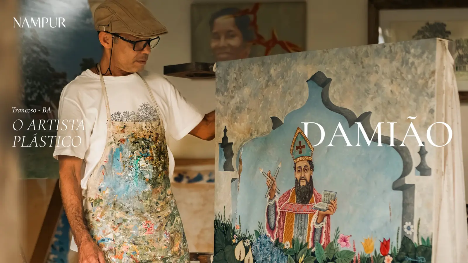 Damião no ateliê, de boina e avental, segurando a bandeira pintada com a imagem de um santo