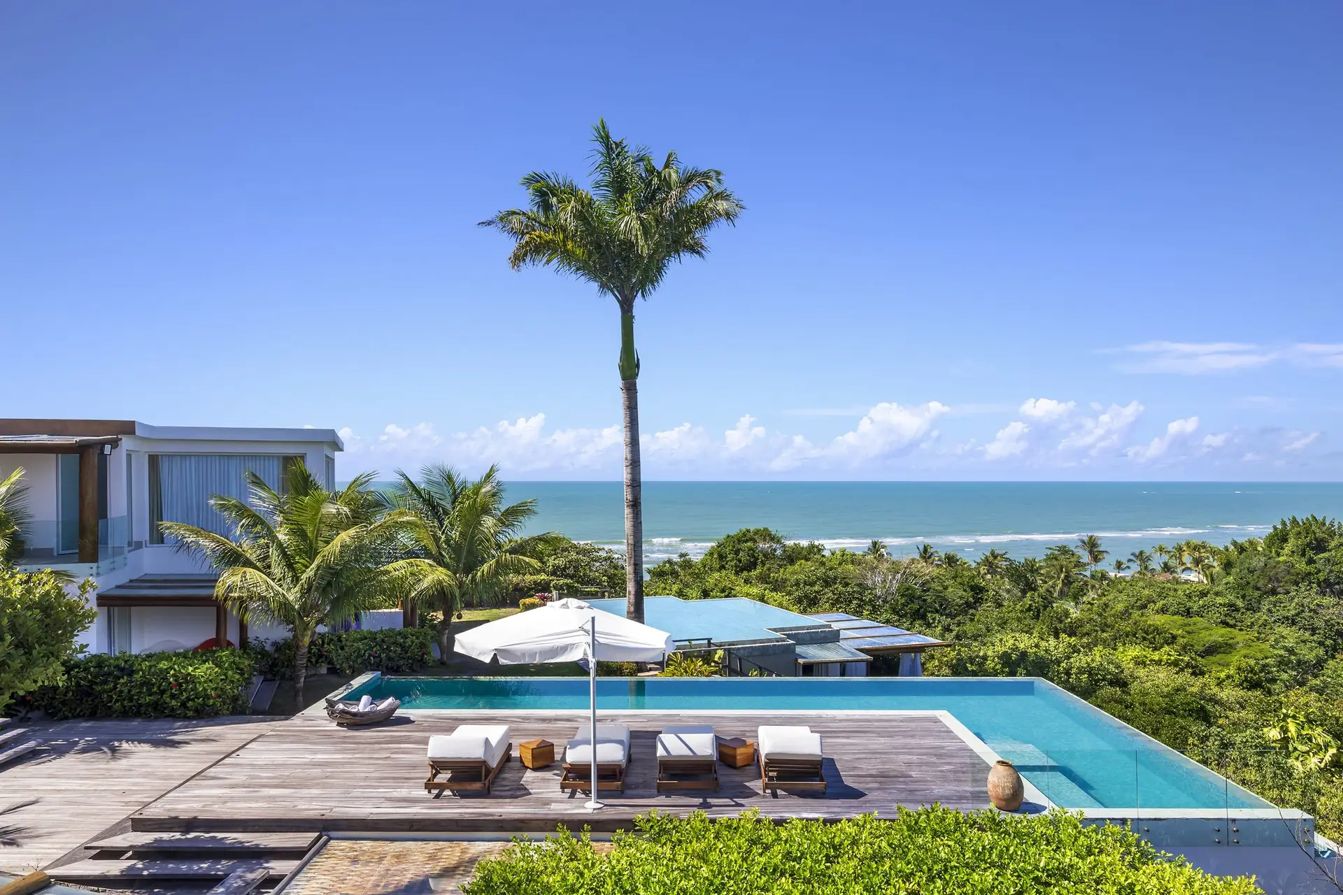 Em um prestigiado retiro costeiro em Trancoso, uma luxuosa piscina infinita oferece vistas deslumbrantes do oceano sob um céu azul claro. O deck de madeira dispõe de três espreguiçadeiras e um amplo guarda-sol branco, todos cercados por altas palmeiras e uma vegetação luxuriante. Este cenário sereno exemplifica o fascínio das casas de praia de alto padrão. Da mesma forma, propriedades impressionantes semelhantes podem ser encontradas nas proximidades de Arraial d'Ajuda.