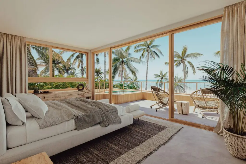 Nestas casas de praia de alto padrão localizadas em Trancoso e Arraial d'Ajuda, um quarto moderno e ensolarado possui uma cama espaçosa adornada com lençóis cinza e complementada por cabeceira de madeira. Um tapete bege adiciona calor ao espaço. Amplas janelas do chão ao teto e portas de correr abrem para uma varanda equipada com duas cadeiras, uma mesa e uma vista encantadora das palmeiras e do mar. Um vaso de planta realça o ambiente aconchegante do cantinho.