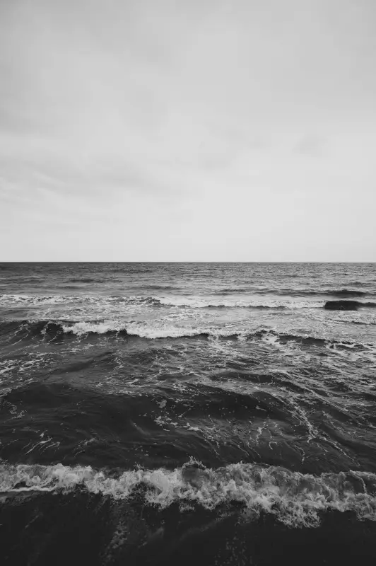 Uma foto em tons de cinza de uma cena serena do oceano captura ondas suaves sob um céu parcialmente nublado que lembra momentos tranquilos nas casas de praia de alto padrão. A imagem retrata a vastidão do mar, com a linha do horizonte quase se fundindo com o céu nublado, evocando lembranças de descanso em Trancoso ou Arraial d'Ajuda.