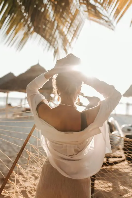 Uma mulher está numa praia perto da sua luxuosa casa de praia, de frente para o sol com as mãos levantadas. Ela está vestida com uma camisa branca e top de biquíni. Folhas de palmeiras e guarda-sóis proporcionam sombra na costa arenosa de Trancoso e Arraial d'Ajuda. A luz do sol lança um brilho quente.