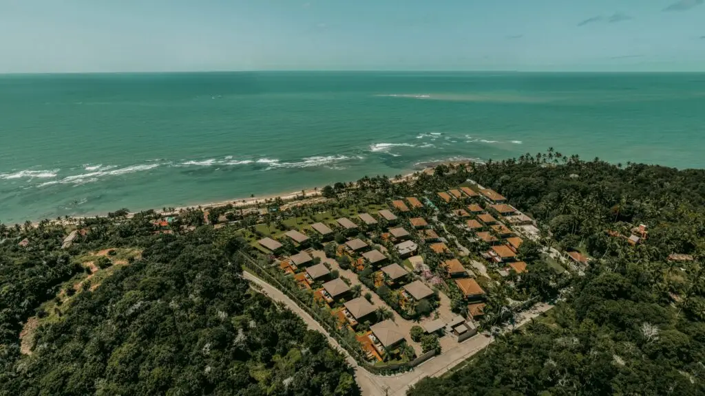 Uma vila de praia de alta qualidade em Trancoso oferece uma vista aérea de uma vila costeira. Fileiras de casas de praia de alto padrão são cercadas por vegetação exuberante. Ao fundo, Arraial d'Ajuda revela o mar azul-turquesa com ondas suavemente encontrando a costa. O céu está quase limpo, pontilhado com algumas nuvens.