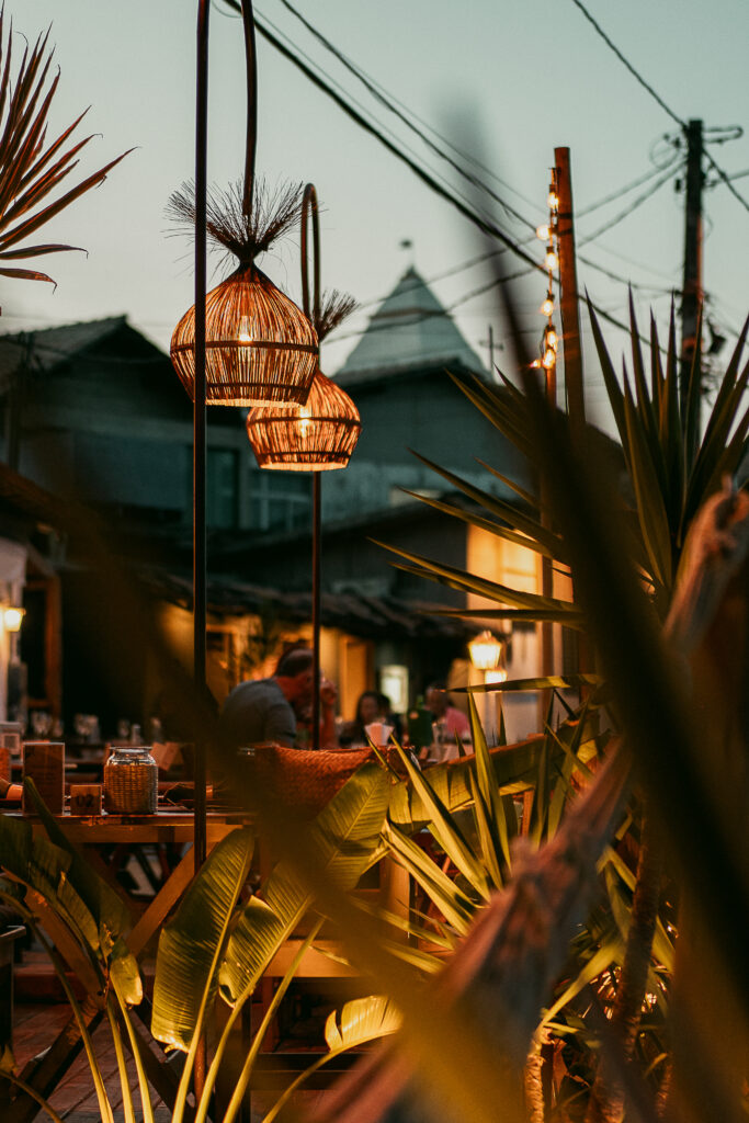 Um cenário de praia de luxo que lembra Trancoso e Arraial d'Ajuda apresenta um café ao ar livre adornado com aconchegantes lâmpadas de vime no teto. Plantas verdes exuberantes realçam o ambiente, enquanto uma mistura de mesas e cadeiras preenche o espaço sob um céu suave da noite, criando uma atmosfera convidativa.