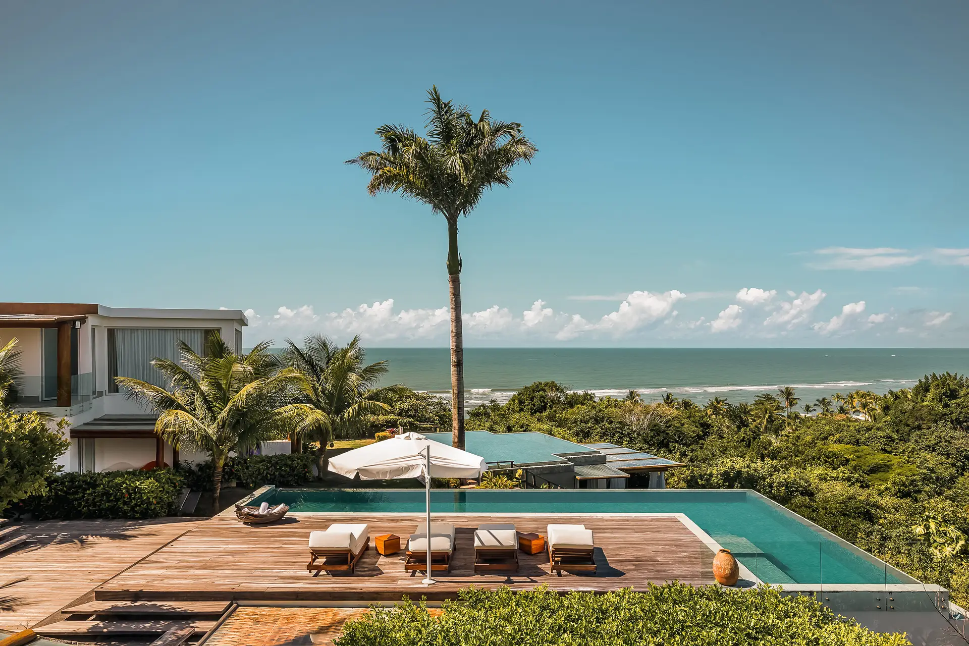 Uma casa de praia de alto padrão em Trancoso exibe uma piscina de borda infinita expansiva e um deck de madeira completo com espreguiçadeiras. Uma palmeira imponente junto com um guarda-sol oferece sombra. A vista abrange vegetação exuberante e o oceano azul claro sob um céu brilhante, típico do cenário deslumbrante em Arraial d'Ajuda.