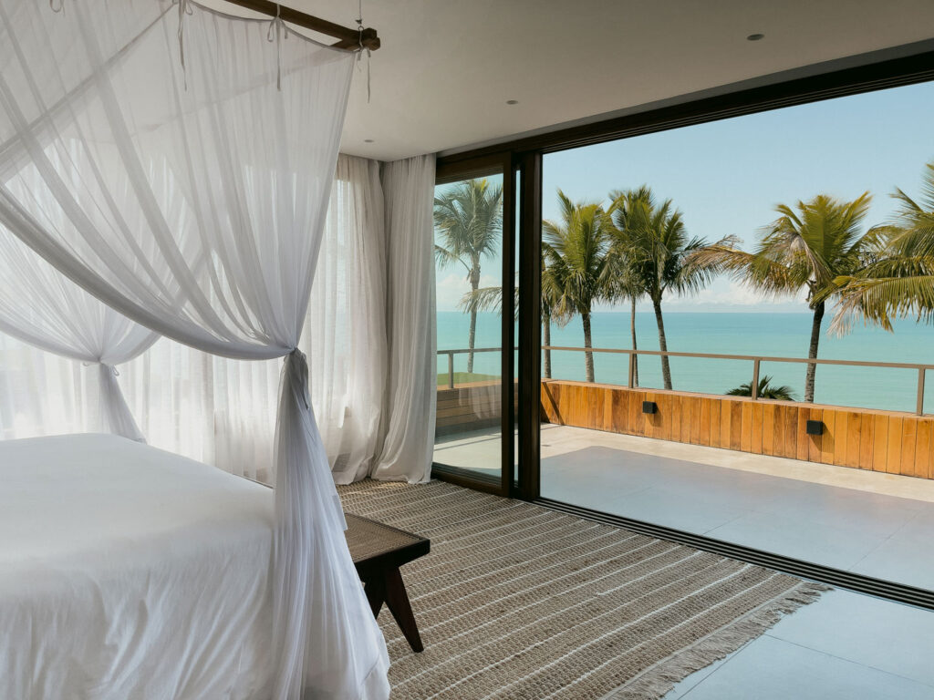 Uma luxuosa casa de praia em Trancoso apresenta um quarto sereno com uma cama de dossel adornada em tecido branco, de frente para uma ampla janela de vidro. A vista pitoresca exibe palmeiras e um oceano calmo sob um céu azul claro, típico das luxuosas casas de praia de alto padrão encontradas em Arraial d'Ajuda. Um tapete texturizado cobre o chão elegantemente.