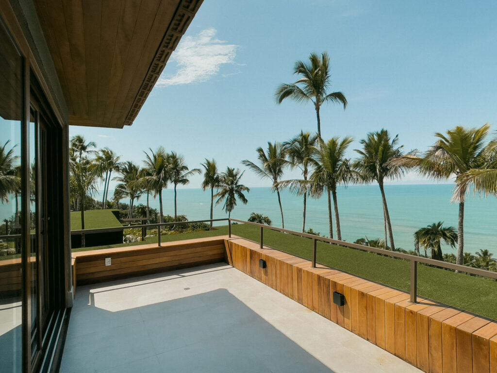 Uma luxuosa varanda de frente para a praia em Trancoso apresenta um corrimão de madeira, oferecendo vistas do pacífico oceano. Palmeiras altas balançam suavemente sob um céu claro, com a luz do sol criando sombras sutis na superfície de azulejos. Esta cena incorpora a vibração tranquila e tropical típica das casas de praia de alto padrão em Arraial d'Ajuda.