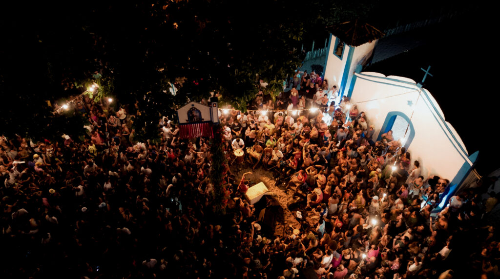 An aerial view captures a festive nighttime gathering near a small, illuminated chapel, surrounded by a crowd holding candles. Above, string lights enhance the atmosphere. Imagine this scene set in areas like Trancoso or Arraial d'Ajuda, known for their luxurious beach houses (casas de praia de alto padrão).
