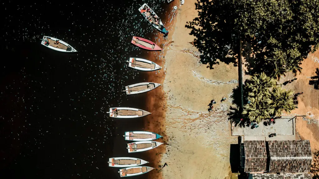 Perspectiva aérea de casas de praia de luxo em Trancoso, perto de Arraial d'Ajuda, revela pequenos barcos alinhados ao longo de um litoral sombrio. A praia de areia faz a transição para uma vegetação exuberante, com palmeiras e um edifício com telhado de palha nas proximidades. Indivíduos são visíveis passeando perto da embarcação.