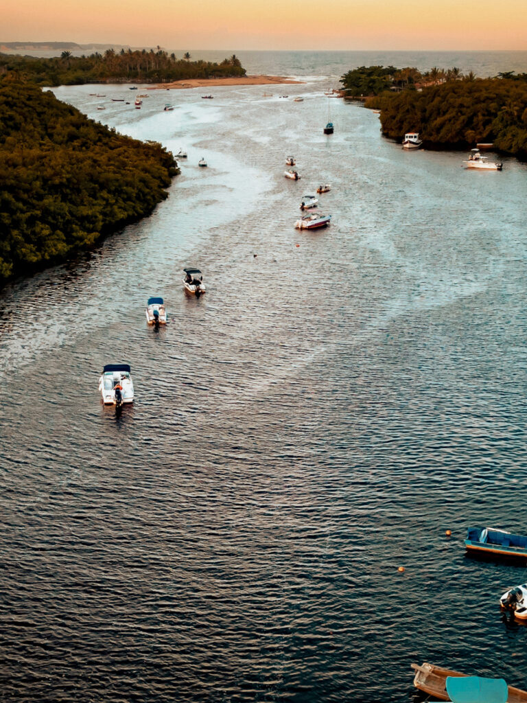 En Trancoso y Arraial d'Ajuda, lujosas casas de playa dan a un río bordeado de árboles verdes y frondosos que se extienden hacia el horizonte. Varias pequeñas embarcaciones están ancladas a lo largo del agua, bajo un cielo pintado de cálidos tonos naranjas y azules, que insinúan el amanecer o el atardecer.