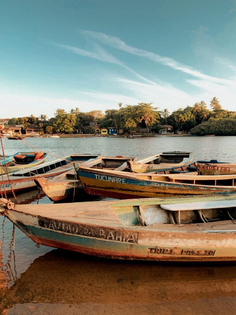 A series of vibrant wooden boats are anchored beside a peaceful riverbank. Beyond them, luxurious beach houses in Trancoso and Arraial d'Ajuda stand amidst verdant foliage under a clear, blue sky.