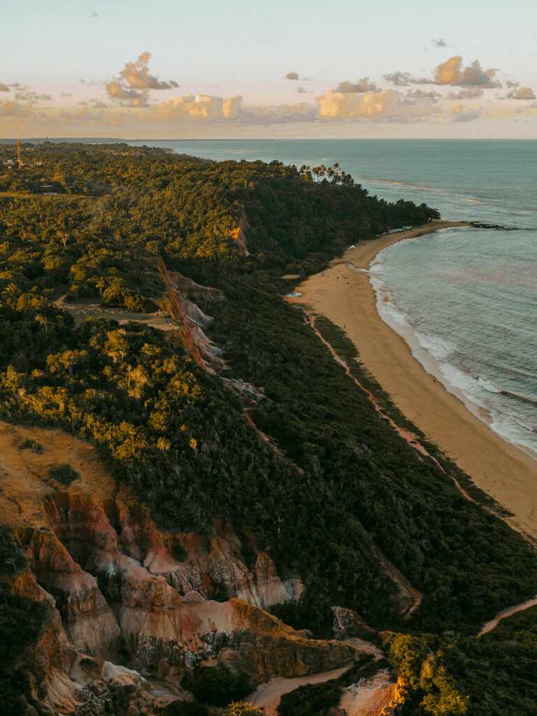 An aerial perspective at sunset showcases the luxurious casas de praia de alto padrão along the coast of Trancoso and Arraial d'Ajuda. A sandy beach is bordered by lush cliffs and a dense forest, while the sea stretches into the distance with scattered clouds overhead.