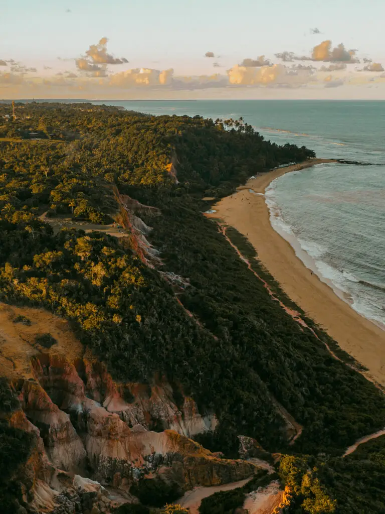 Uma perspectiva aérea revela o litoral de Trancoso e Arraial d'Ajuda ao pôr do sol, com casas de praia de alto padrão. Uma longa praia de areia se estende ao longo de penhascos verdejantes com vegetação. O oceano calmo reflete os tons dourados do céu, enquanto nuvens dispersas enriquecem o ambiente quente e sereno.