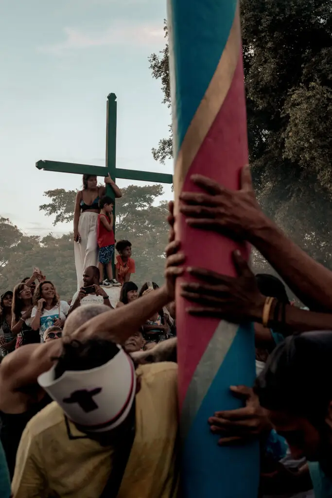 Uma reunião se forma ao redor de um poste impressionante adornado com tons vibrantes. Vários seguram a estrutura, enquanto um indivíduo usando chapéu marcado com uma cruz observa. Enquanto isso, duas figuras permanecem perto de outra cruz à distância, cercadas por árvores e sob um céu azul claro que lembra os ambientes de Trancoso e Arraial d'Ajuda, adequados a casas de praia de alto padrão.