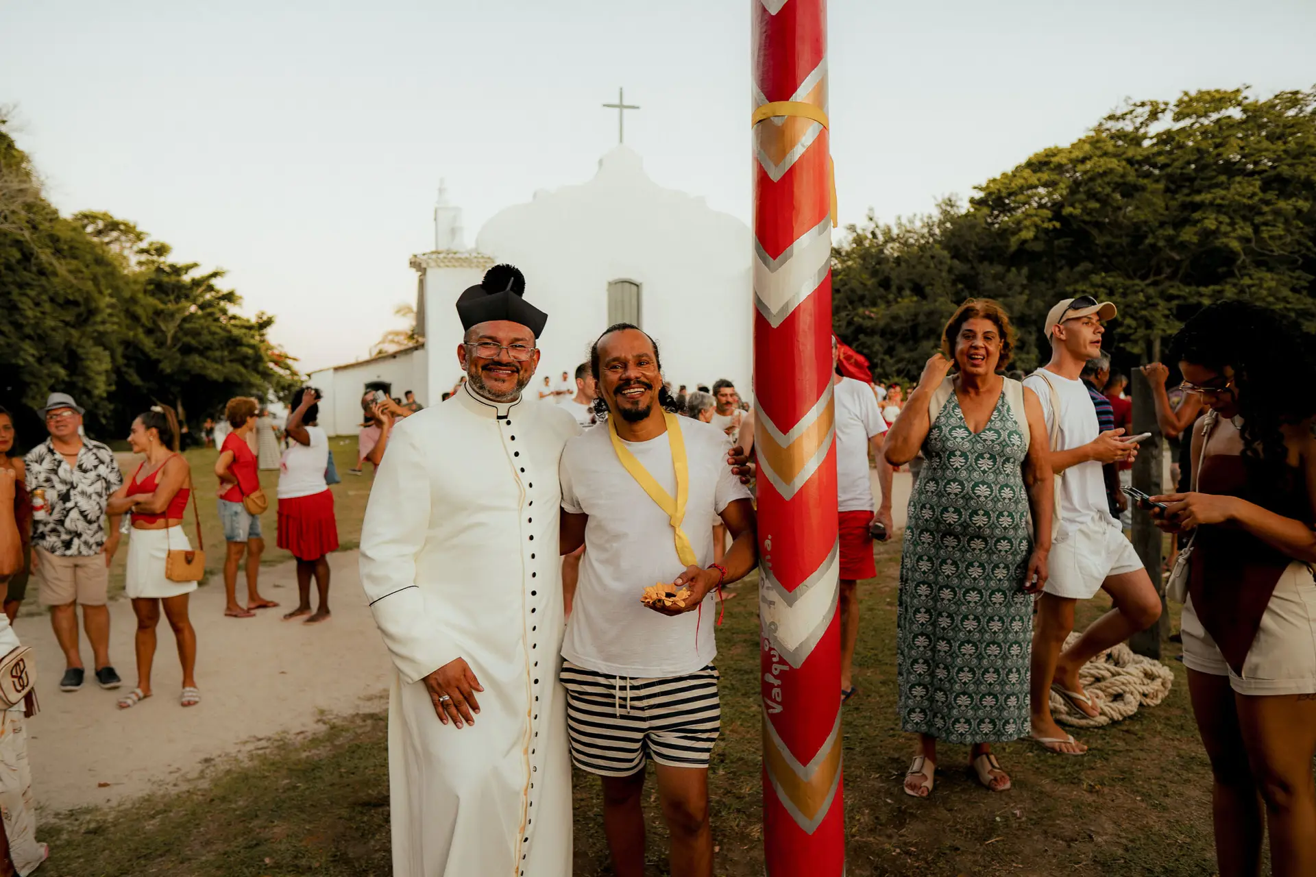 Em um ambiente animado ao ar livre, perto de uma pitoresca igreja branca encimada por uma cruz, em meio a uma multidão festiva, um indivíduo veste uma túnica religiosa e chapéu, enquanto outro ostenta listras e um cachecol amarelo. Esta cena vibrante lembra as casas de praia de alto padrão encontradas em Trancoso e Arraial d'Ajuda.