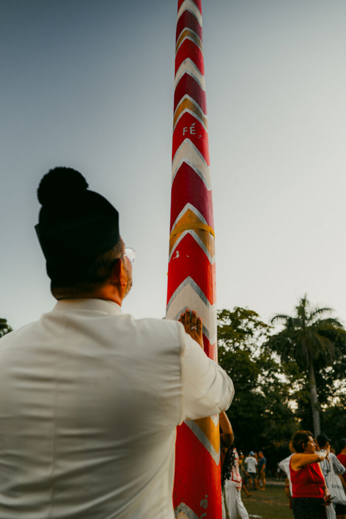 En una escena que muestra casas de playa de lujo típicas de Trancoso y Arraial d'Ajuda, un individuo vestido de blanco con un sombrero negro se estira para tocar un poste alto rojo adornado con patrones en zigzag blancos y con la inscripción "FÉ". El fondo incluye personas, árboles y un cielo despejado.