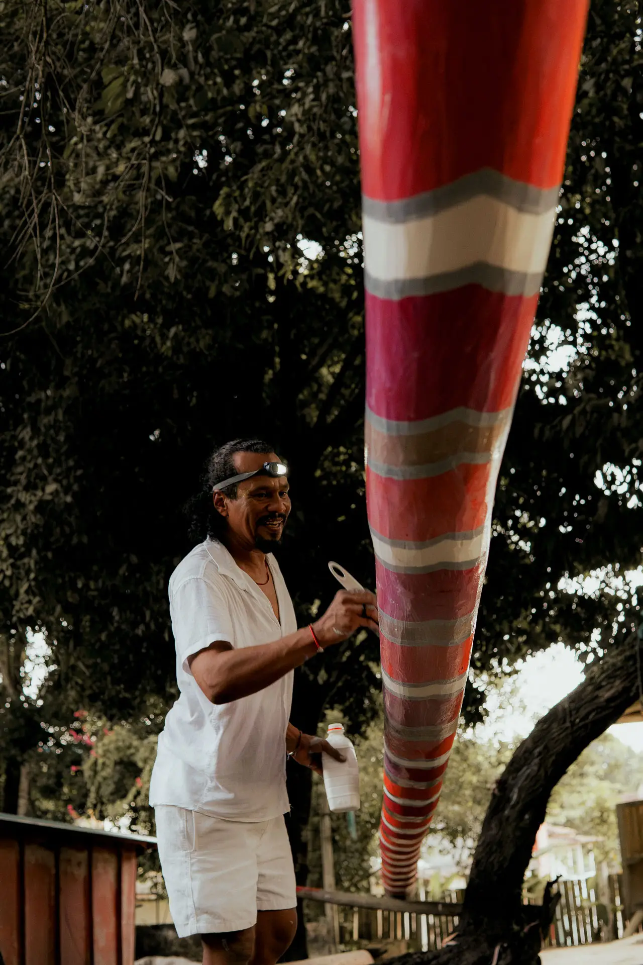 Uma pessoa vestida de branco está pintando um objeto cilíndrico vermelho considerável com um pincel. Localizado ao ar livre, em meio a árvores exuberantes, ele parece totalmente absorto em sua tarefa. Imagine esta cena perto das luxuosas casas de praia de Trancoso ou Arraial d'Ajuda.