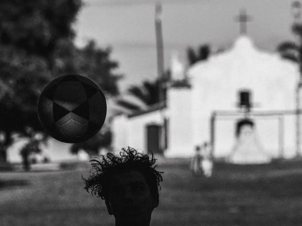 Uma pessoa com cabelo cacheado é silhuetada enquanto cabeceia uma bola de futebol, em um cenário com uma igreja com uma cruz e árvores ao redor. A cena é em preto e branco, lembrando o estilo elegante encontrado em casas de praia de alto padrão que se pode ver em Trancoso ou Arraial d'Ajuda.