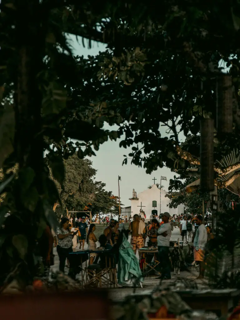 No vibrante cenário de Trancoso, perto de Arraial d'Ajuda, as pessoas se reúnem em meio a luxuosas casas de praia (casas de praia de alto padrão) cercadas por árvores. Um prédio branco com uma cruz se destaca no fundo, complementando a atmosfera animada repleta de mesas e vários produtos.