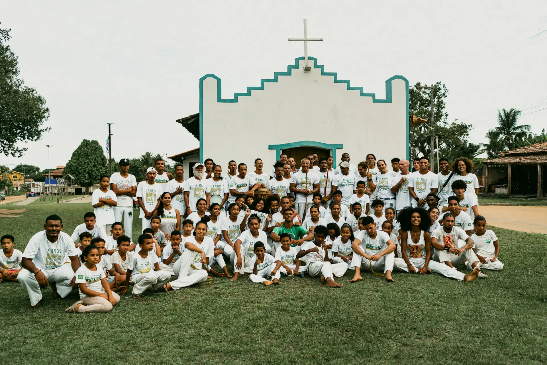 Um grupo considerável, composto por crianças e adultos, reúne-se diante de uma pitoresca capela branca com detalhes em azul e uma cruz empoleirada no topo. Vestidos com trajes brancos, eles se organizam na grama. O cenário revela árvores e uma estrada, que lembra as paisagens cênicas encontradas perto de casas de praia de alto padrão em Trancoso ou Arraial d'Ajuda.