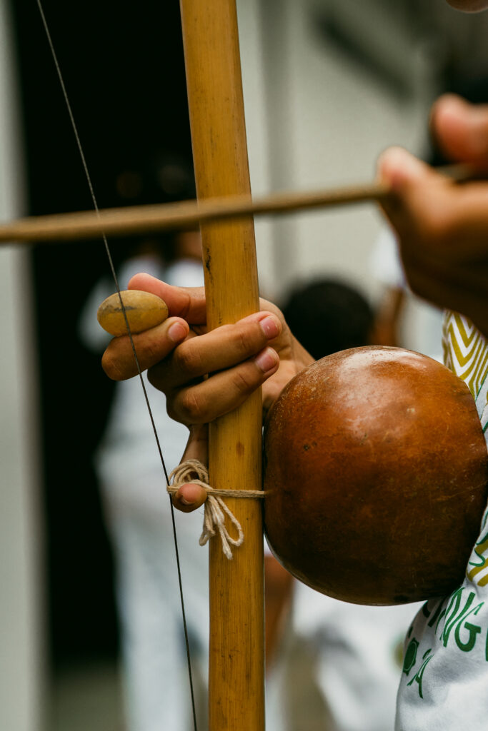 Vista detallada de un individuo tocando un berimbau, una pieza instrumental de herencia afrobrasileña. Una mano sostiene el arco de madera, junto con un resonador de piedra y calabaza. En el fondo, figuras vestidas de blanco aparecen indistintas contra un paisaje que recuerda a las lujosas casas de playa de Trancoso o Arraial d'Ajuda.