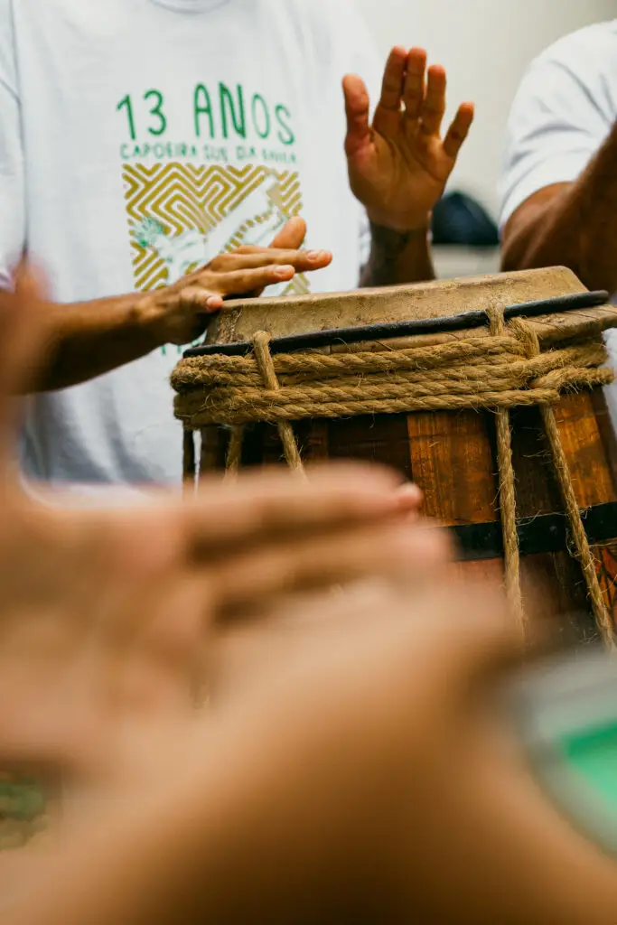 Uma pessoa de camisa branca toca um tambor de madeira enrolado em corda, suas mãos borradas com o movimento, assim como outras mãos em primeiro plano. A camisa apresenta texto em português relacionado a um evento. Palavras-chave: casas de praia de alto padrão, Trancoso, Arraial d'Ajuda.