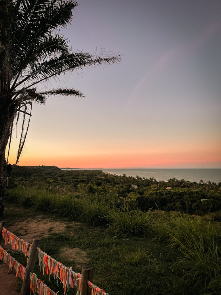 Une vue pittoresque sur les maisons de plage haut de gamme de Trancoso et d'Arraial d'Ajuda présente un premier plan herbeux avec un palmier positionné à gauche. Le ciel affiche des nuances de rose et d'orange, projetant une lueur sereine sur la mer tranquille et l'horizon lointain. Une clôture ornée de rubans colorés renforce le charme de la scène.