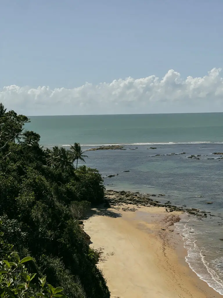 Um cenário pitoresco de litoral apresenta luxuosas casas de praia de alto padrão em Trancoso e Arraial d'Ajuda. A praia de areia encontra águas calmas do oceano azul-esverdeado sob um céu adornado com nuvens fofas. Árvores verdejantes e pedras espalhadas margeiam a costa, completando esta paisagem deslumbrante.