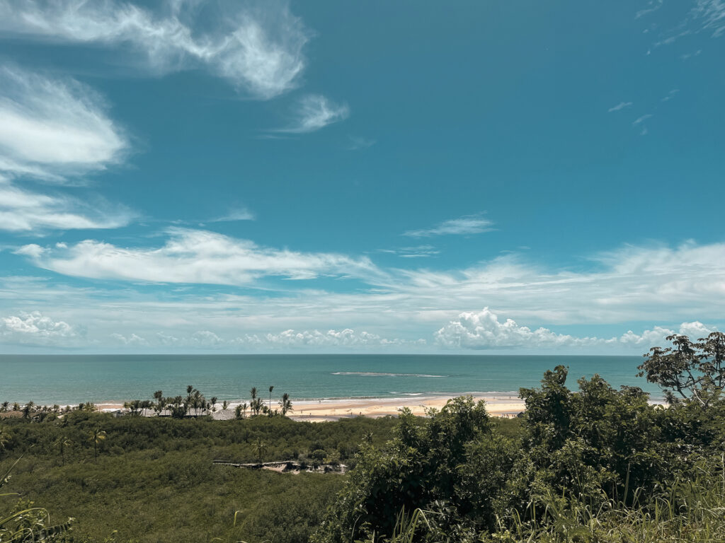 Una vista impresionante muestra las casas de playa de lujo en Trancoso y Arraial d'Ajuda. La vegetación exuberante llena el primer plano, mientras que a lo lejos, el océano besa la costa arenosa bajo un cielo despejado adornado con nubes dispersas. Algunas palmeras se suman al pintoresco entorno.