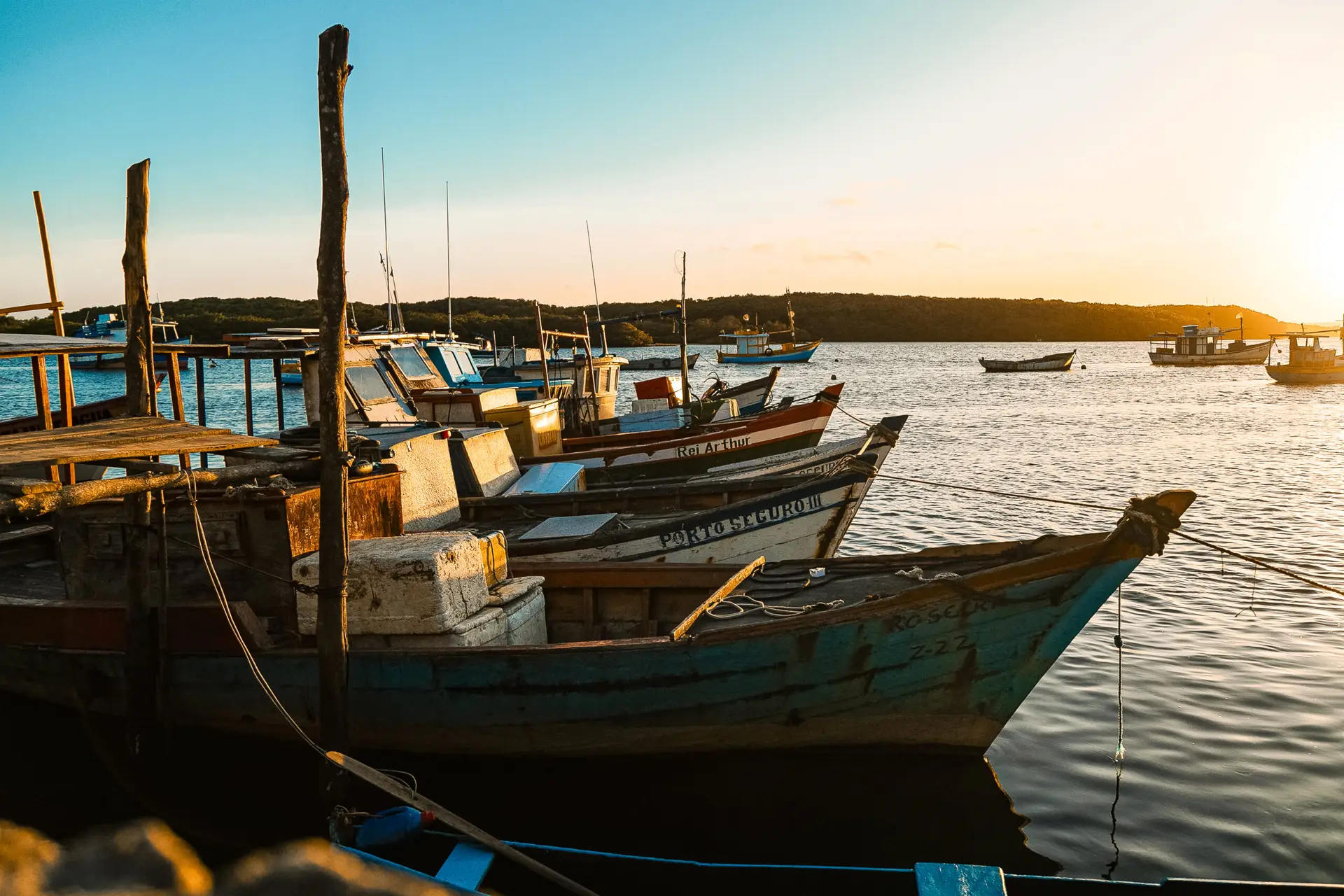 Casas de praia de alto padrão em Trancoso e Arraial d'Ajuda refletem o pôr do sol dourado sobre águas calmas, enquanto barcos de madeira estão próximos ao píer, criando uma paisagem serena e sofisticada.