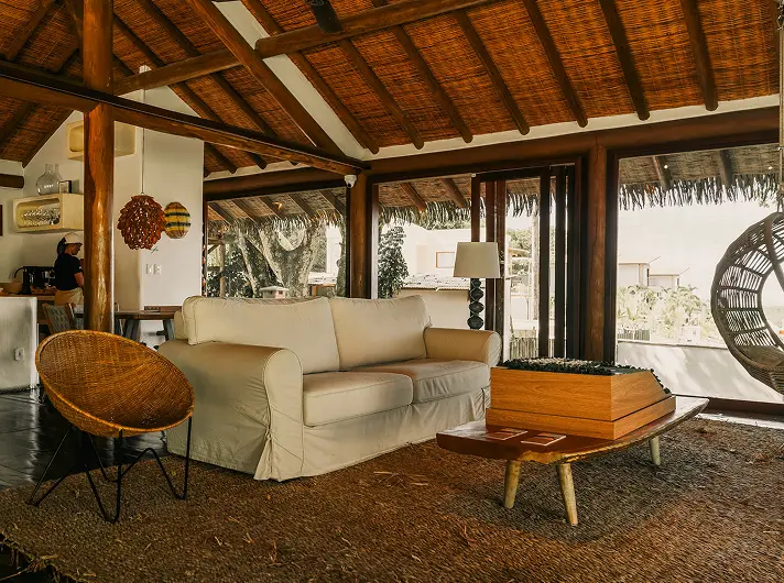 Refined beach house in Trancoso or Arraial d'Ajuda: spacious living area with sunlit ambiance, white sofa, woven chairs, wooden coffee table, expansive rug, exposed beams overhead, and large windows offering views to a patio surrounded by lush greenery—exemplifying casas de praia de alto padrão.