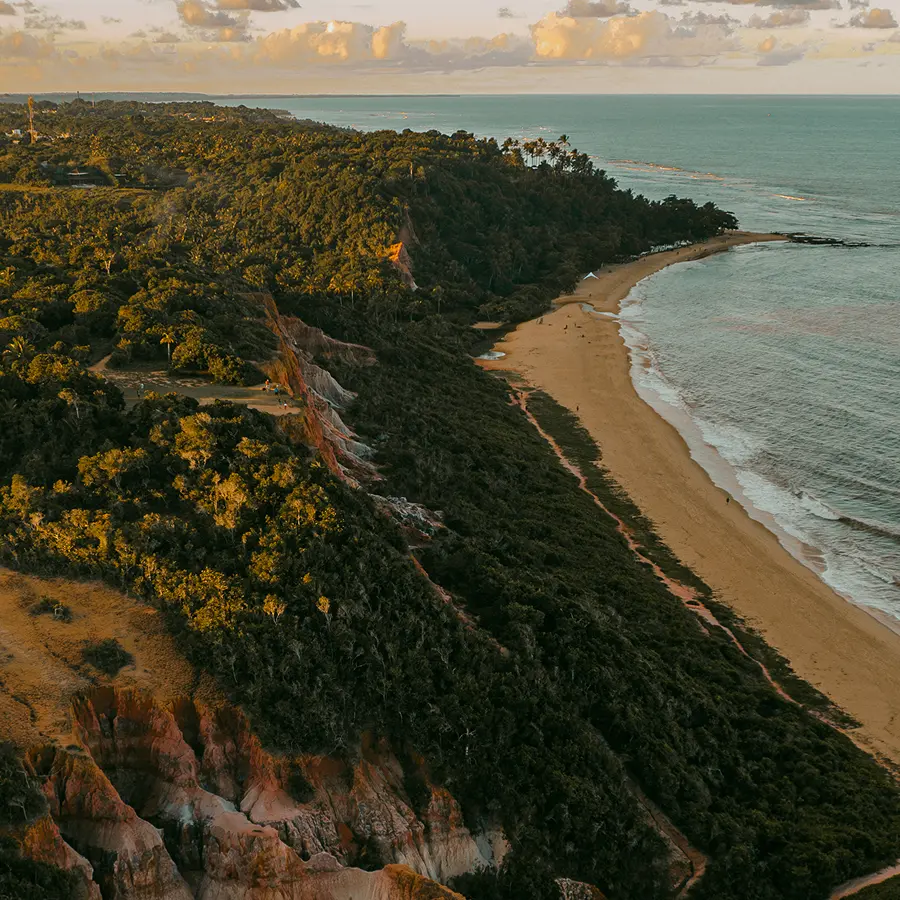 Vista aérea das casas de praia de alto padrão situadas ao longo da costa entre Trancoso e Arraial d'Ajuda, com praias de areia, ondas do mar e falésias verdejantes sob um céu parcialmente nublado ao pôr do sol.
