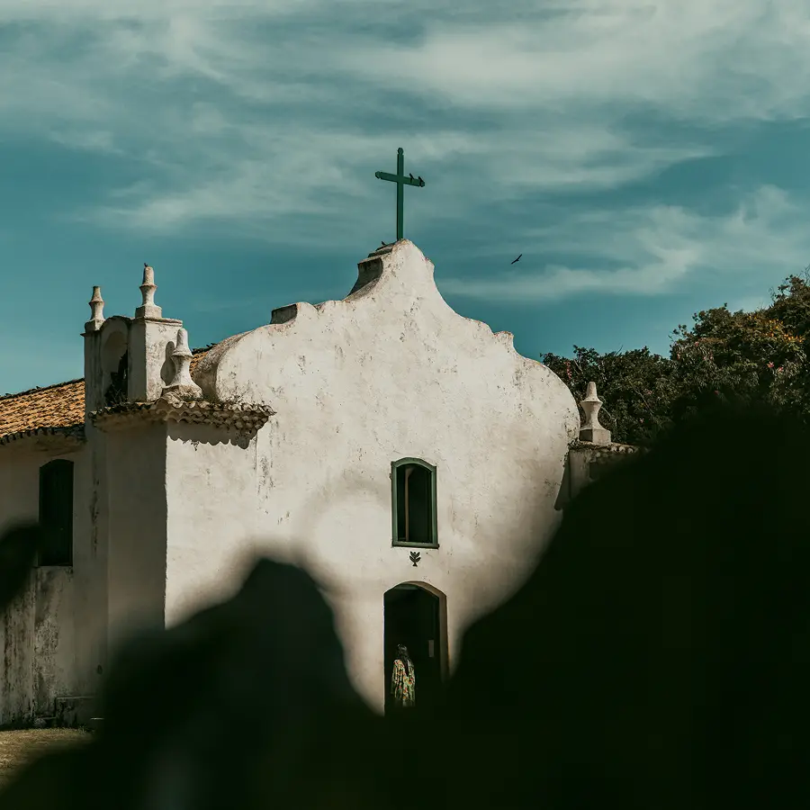 Casa de praia de alto padrão em Trancoso, com elementos que lembram uma pequena igreja branca com uma cruz verde no topo do telhado, emoldurada por uma densa folhagem em primeiro plano. A estrutura rústica apresenta um charme do velho mundo contra um céu azul parcialmente nublado, cercado por árvores - capturando a atmosfera exclusiva encontrada nas casas de praia de alto padrão típicas de Trancoso e Arraial d'Ajuda.