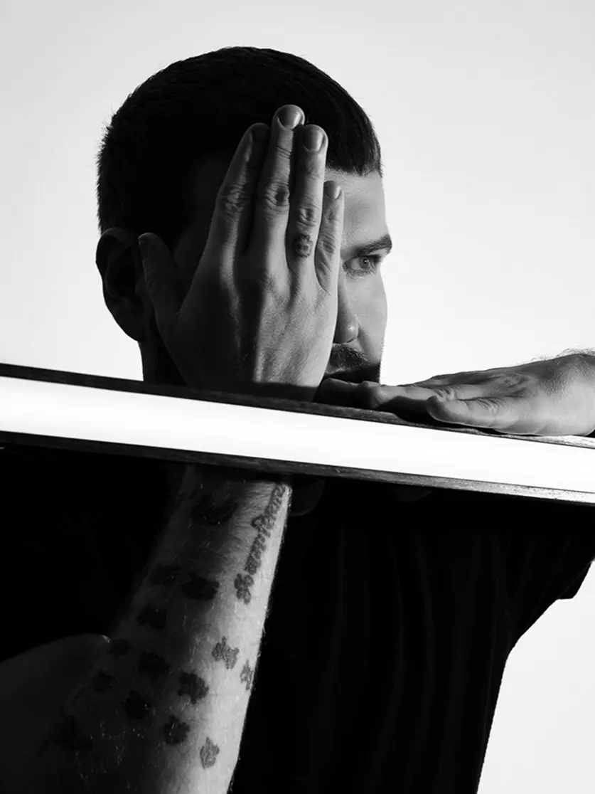 Uma imagem em preto e branco mostra um homem posando com um olho coberto e o braço apoiado em uma faixa de luz horizontal; a cena evoca o ambiente sofisticado encontrado em casas de praia de alto padrão típicas de Trancoso e Arraial d'Ajuda.