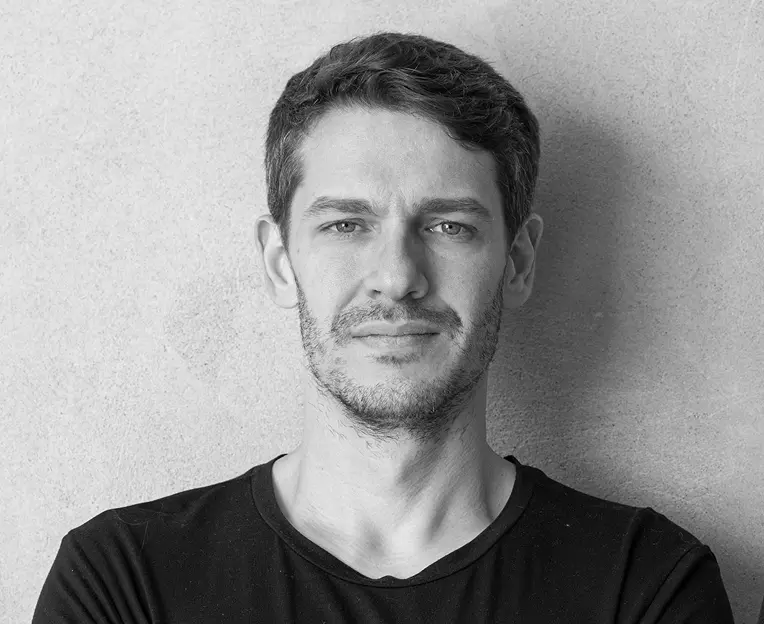 Uma imagem em preto e branco mostra um homem com cabelos escuros curtos e barba aparada, vestindo uma camisa simples, de pé contra uma parede texturizada. A cena evoca o estilo refinado das casas de praia de alto padrão encontradas em Trancoso e Arraial d'Ajuda.