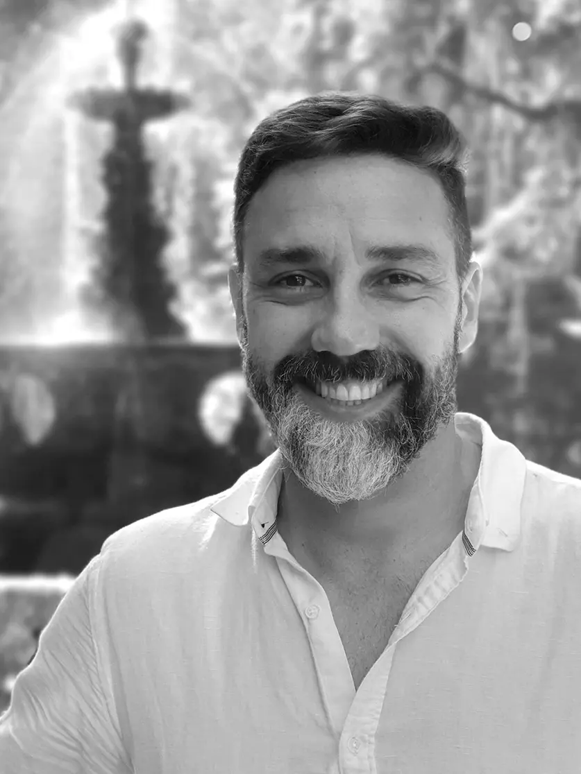 Um homem com barba e cabelo curto, vestido com uma camisa clara de botões, sorri para a câmera em uma fotografia em preto e branco. Atrás dele, a luz do sol passa por entre as árvores e destaca uma fonte de jardim embaçada - evocando o ambiente sofisticado das casas de praia de alto padrão, normalmente encontradas em Trancoso e Arraial d'Ajuda.