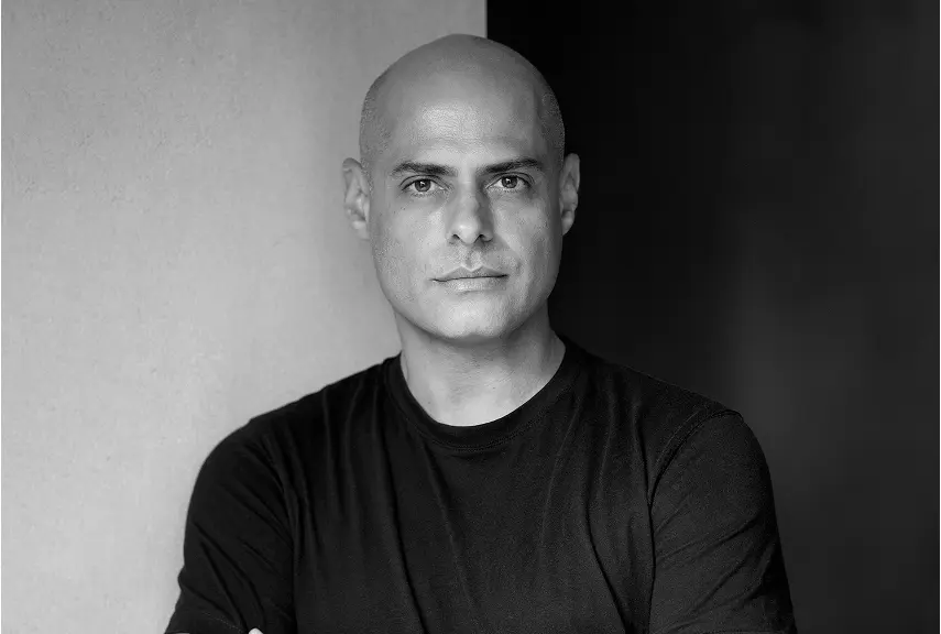 Um homem está de braços cruzados, olhando para a câmera, em um cenário contrastante de luz e escuridão. A imagem em preto e branco evoca o ambiente sofisticado das casas de praia de alto padrão encontradas em Trancoso e Arraial d'Ajuda.