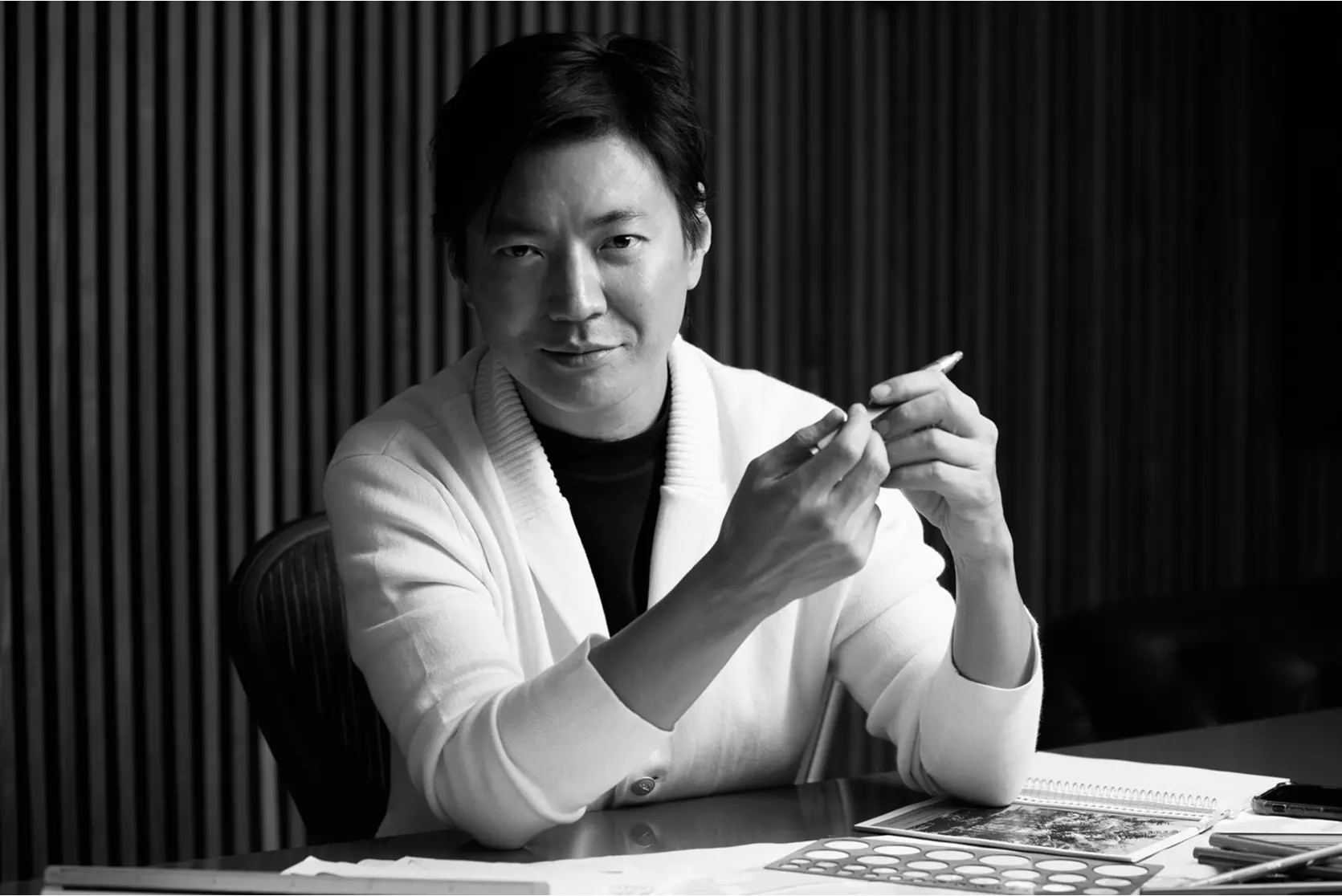 Em uma foto em preto e branco com um fundo de madeira listrada, um homem está sentado em uma mesa com papéis e caderno, com uma caneta na mão, olhando para a câmera - um ambiente que lembra as casas de praia de alto padrão encontradas em Trancoso e Arraial d'Ajuda.