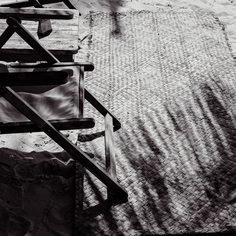 Cena em preto e branco de uma esteira trançada na areia, a luz do sol projetando a sombra de uma cadeira, evocando casas de praia de alto padrão em Trancoso, Arraial d'Ajuda.