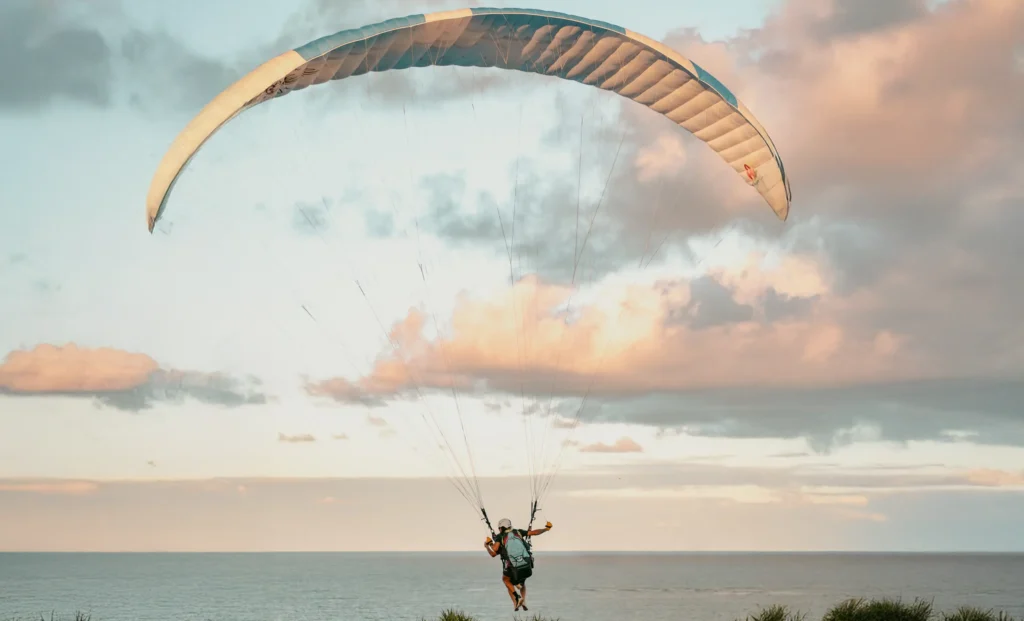 Paraglider soars above grassy terrain near casas de praia de alto padrão in Trancoso and Arraial d'Ajuda at sunset, pastel sky above.