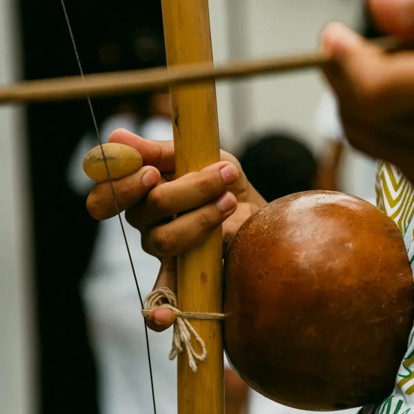 A detailed view captures an individual interacting with a berimbau, a traditional Brazilian string instrument. Hands are seen manipulating a stick and gourd, while clad in a white shirt. The scene's backdrop is softly blurred. Imagine this artistic portrayal amidst the luxurious beach houses of Trancoso or Arraial d'Ajuda's high-end coastal residences.