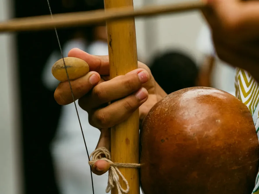 A detailed view captures an individual interacting with a berimbau, a traditional Brazilian string instrument. Hands are seen manipulating a stick and gourd, while clad in a white shirt. The scene's backdrop is softly blurred. Imagine this artistic portrayal amidst the luxurious beach houses of Trancoso or Arraial d'Ajuda's high-end coastal residences.