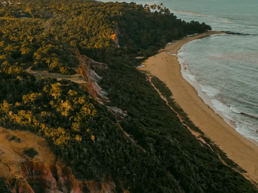 An aerial perspective at sunset showcases the luxurious casas de praia de alto padrão along the coast of Trancoso and Arraial d'Ajuda. A sandy beach is bordered by lush cliffs and a dense forest, while the sea stretches into the distance with scattered clouds overhead.