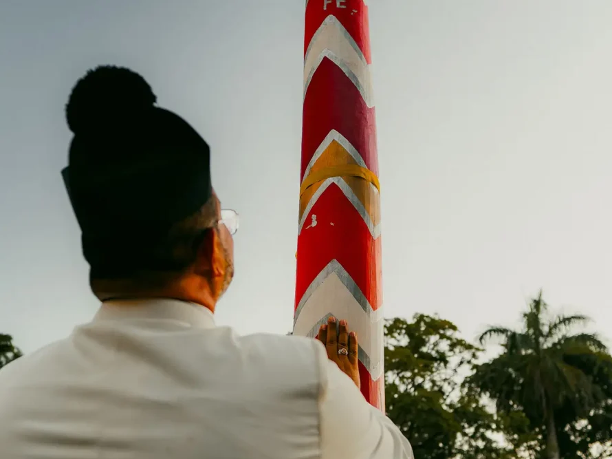 Dressed in a white outfit and black cap, a person touches a tall red and white decorated pole outdoors. The clear sky reveals trees and individuals in the background, reminiscent of the scenic beauty found around high-end beach houses like those in Trancoso or Arraial d'Ajuda.
